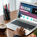 Laptop mit geöffneter Webdesign-Website auf einem Schreibtisch neben Stiften und Kaffee