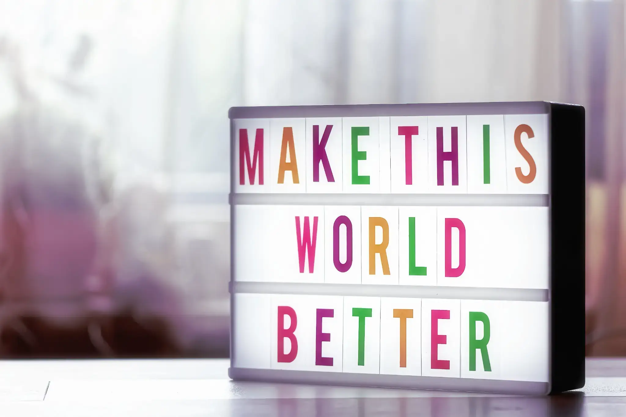 Bunter Leuchtkasten mit dem englischen Motivationsspruch „Make This World Better“ auf einem Tisch